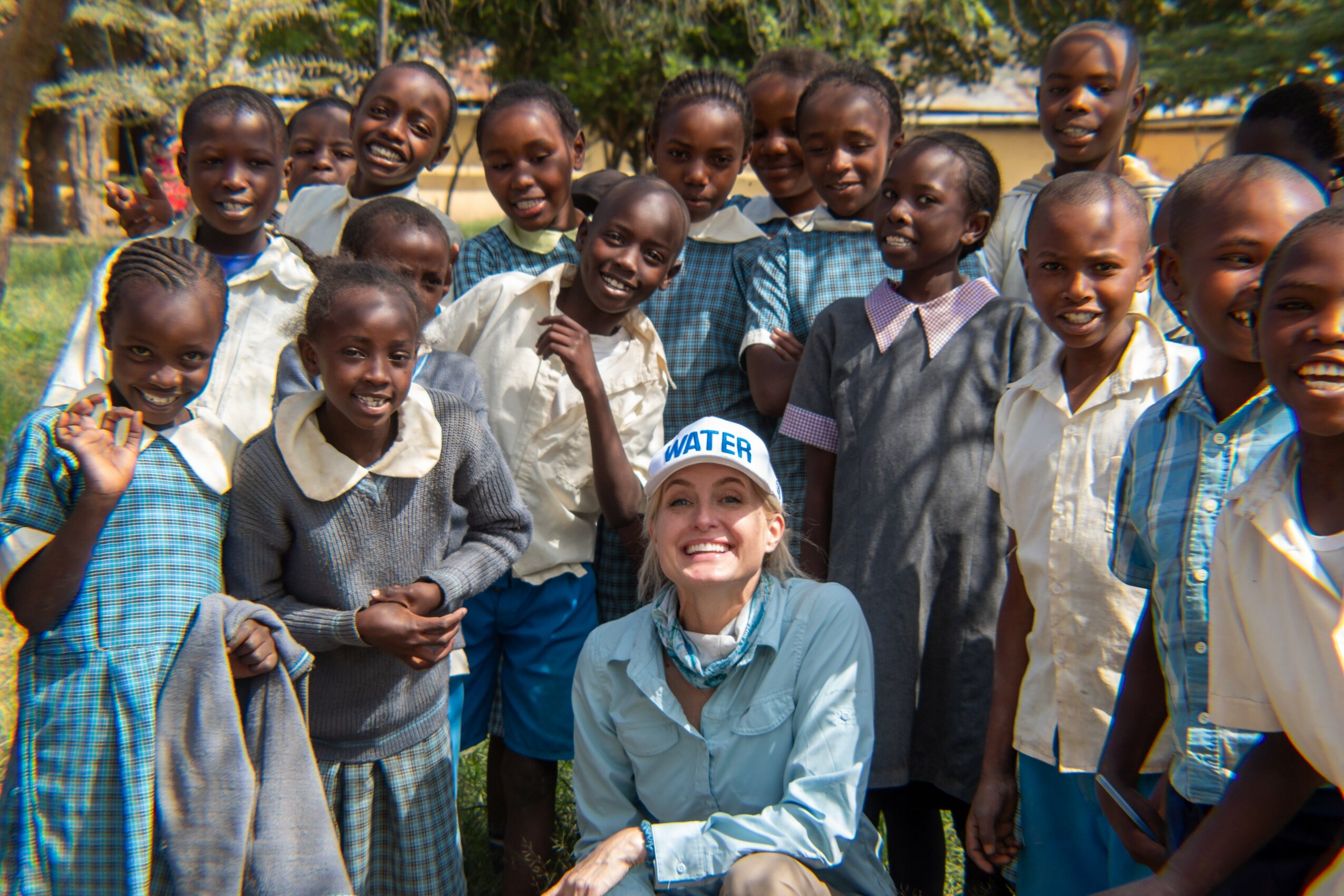 Group of children and a woman outdoors, smiling, with trees in background.