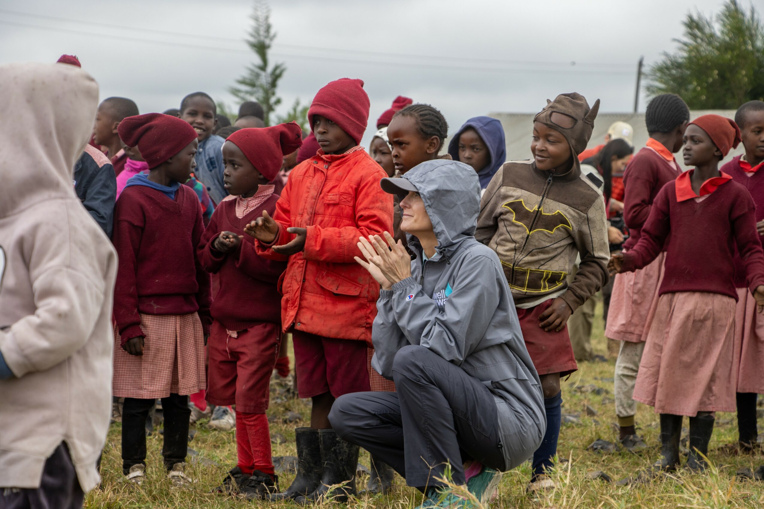 Group of children and an adult outdoors, some children wearing red and pink clothing, with cloudy sky in background.