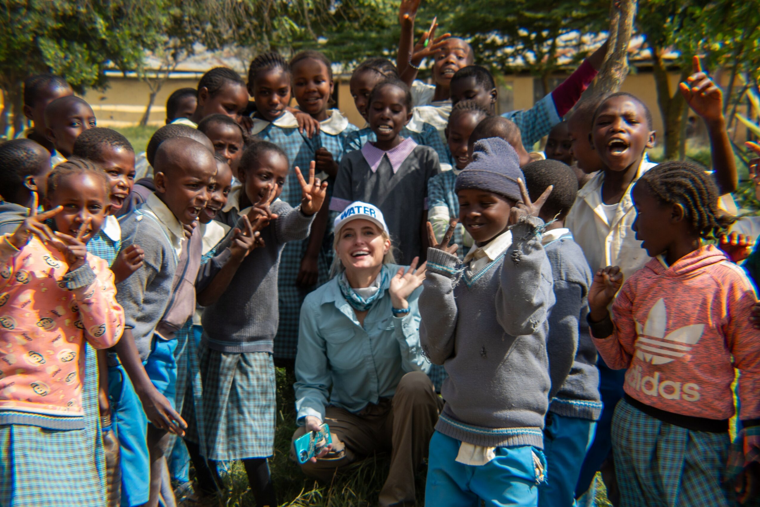 Group of children and a woman outdoors, smiling and waving, with trees in the background.
