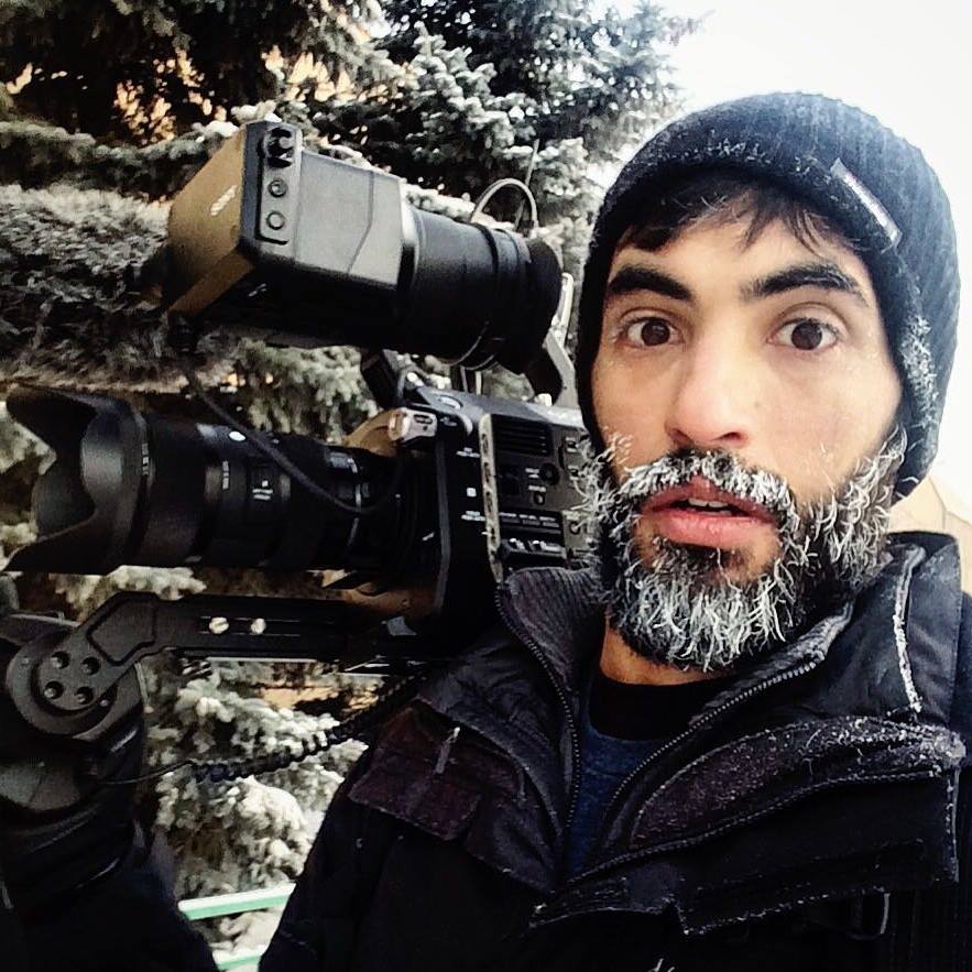 Man with a beard and black beanie holding a professional camera outdoors in winter, snow-covered trees in background.