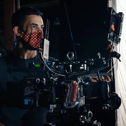 Person operating a camera with a red patterned face mask, in a dark environment, surrounded by equipment.