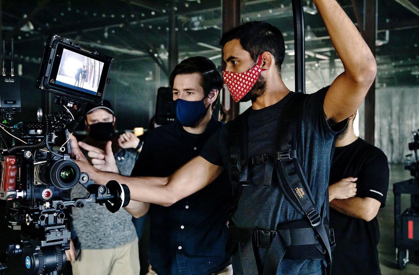 Group of people wearing masks, operating a professional camera indoors, with glass windows and reflections in the background.
