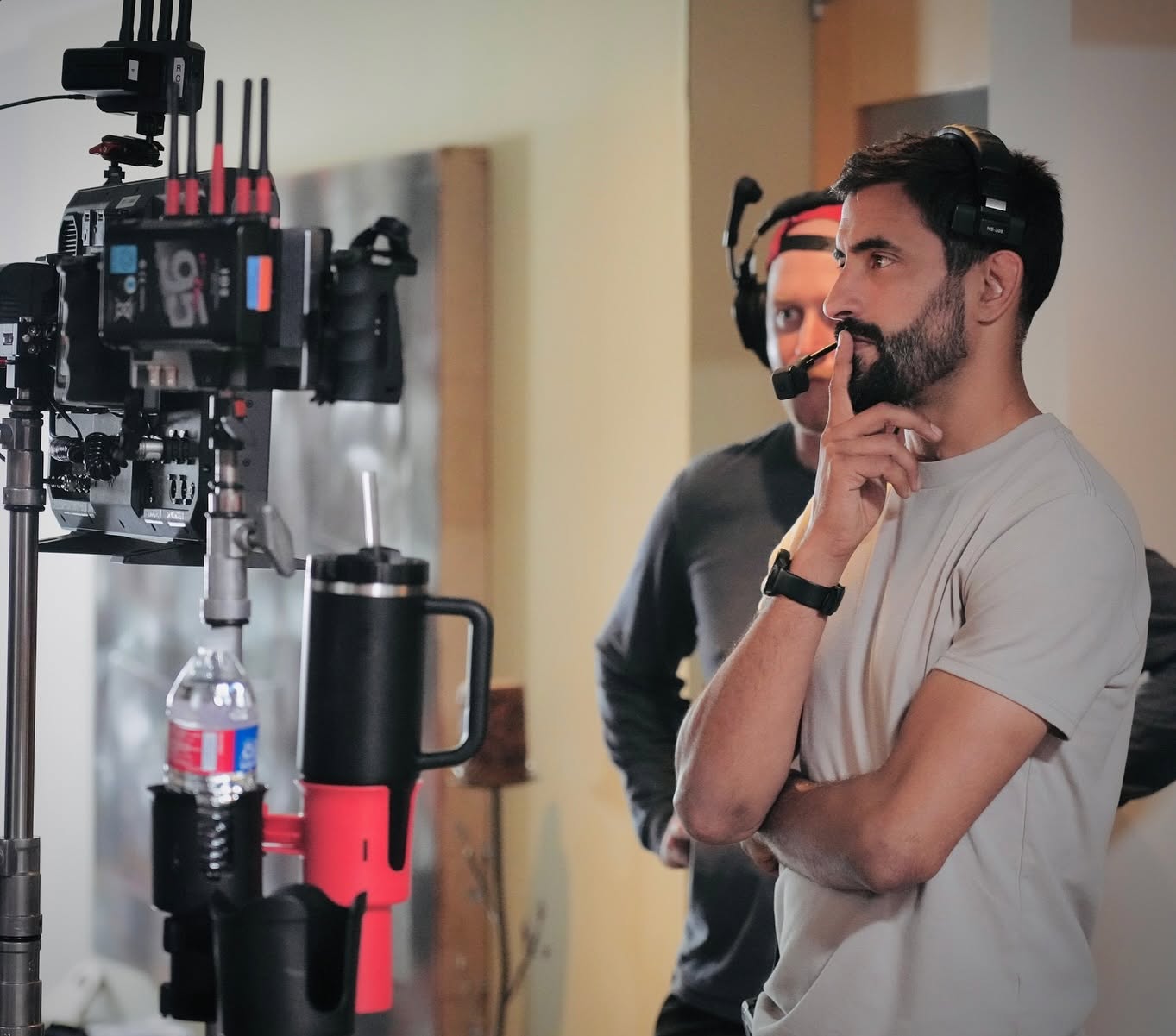 Two men stand near camera equipment, one with a beard and white shirt, the other with headphones, smiling.
