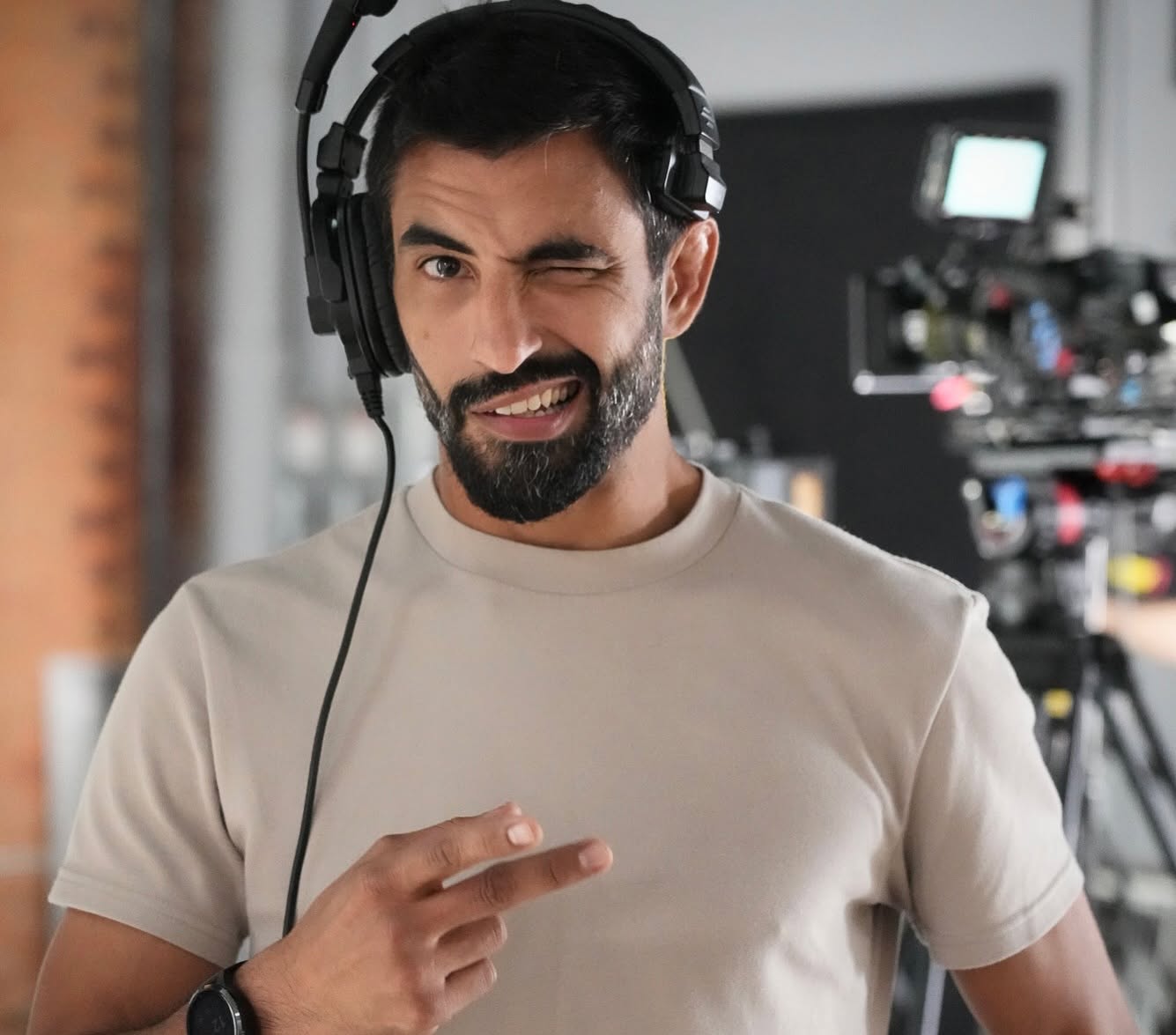 Man with a beard wearing headphones, pointing at himself, in a studio with camera equipment in the background.