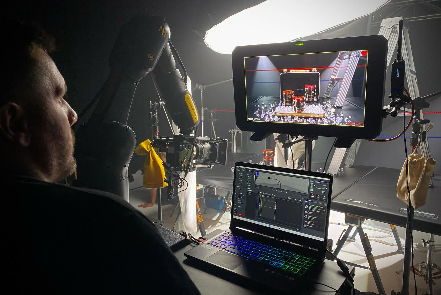 Person operating camera equipment with monitors and a laptop in a studio setting.
