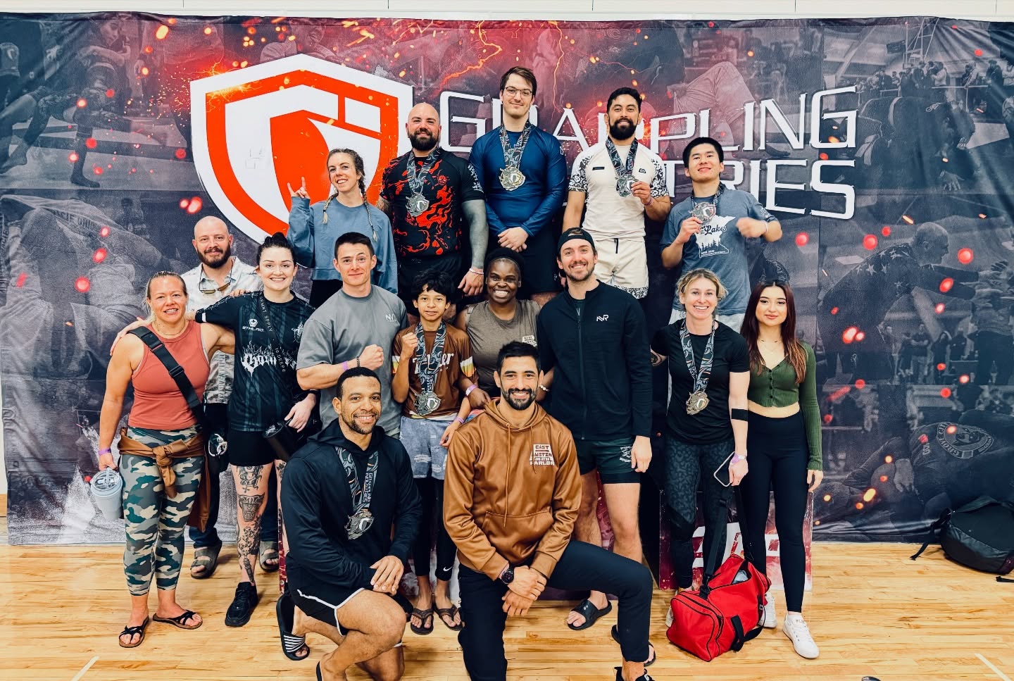Group of 15 people standing and kneeling in front of a gaming event backdrop with a shield logo, smiling.