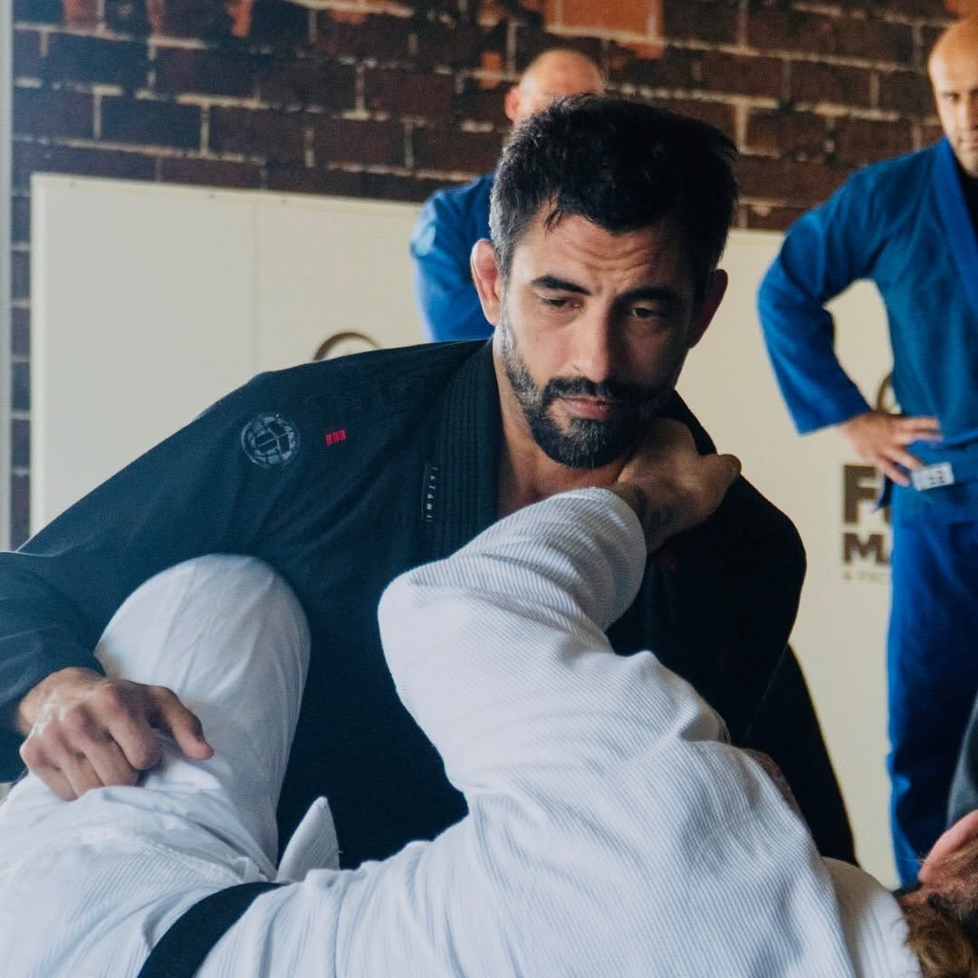 A man in a black martial arts uniform with a beard looks down at a person in a white gi, with others in blue uniforms nearby.