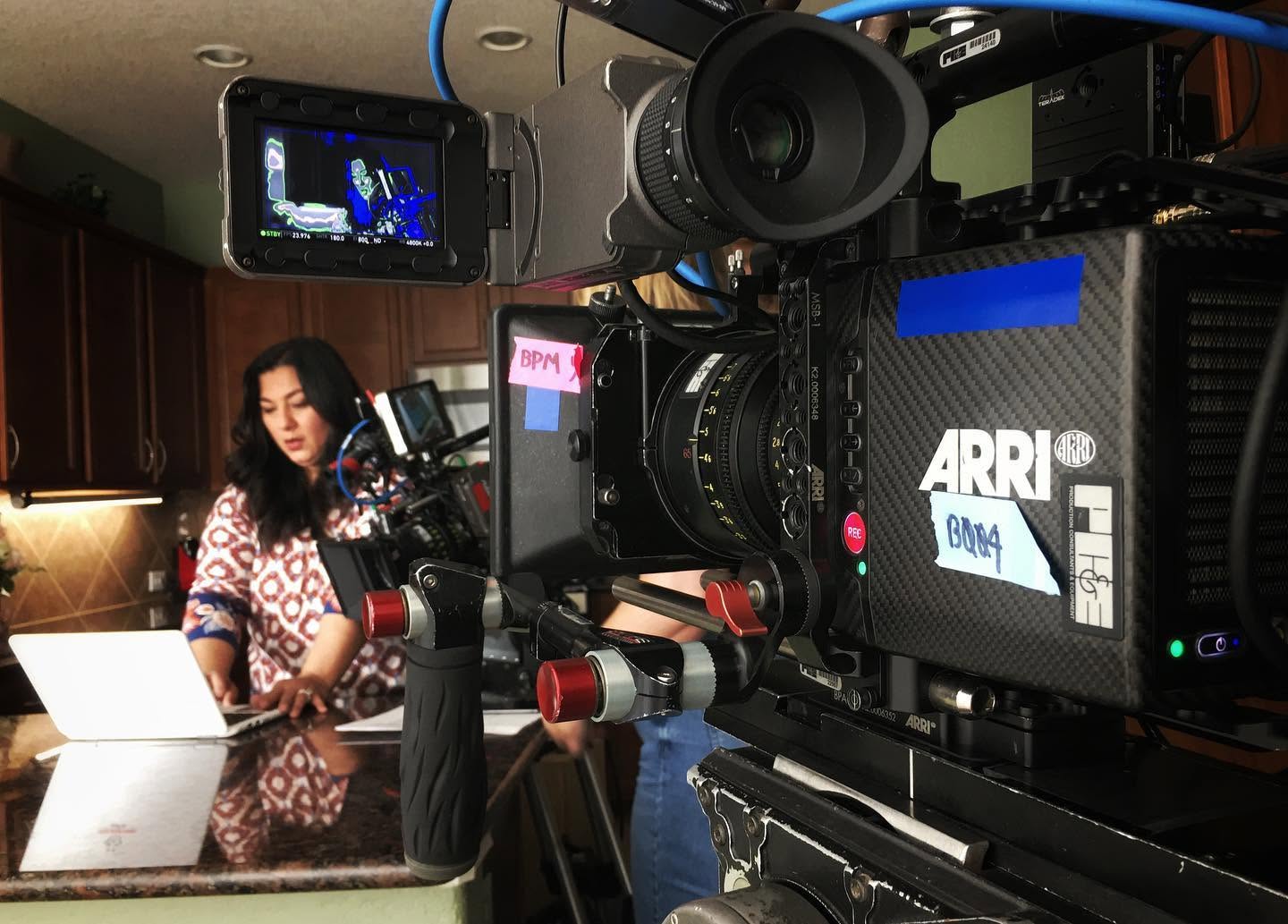 Woman in patterned shirt working in a kitchen studio with camera equipment and monitors nearby.