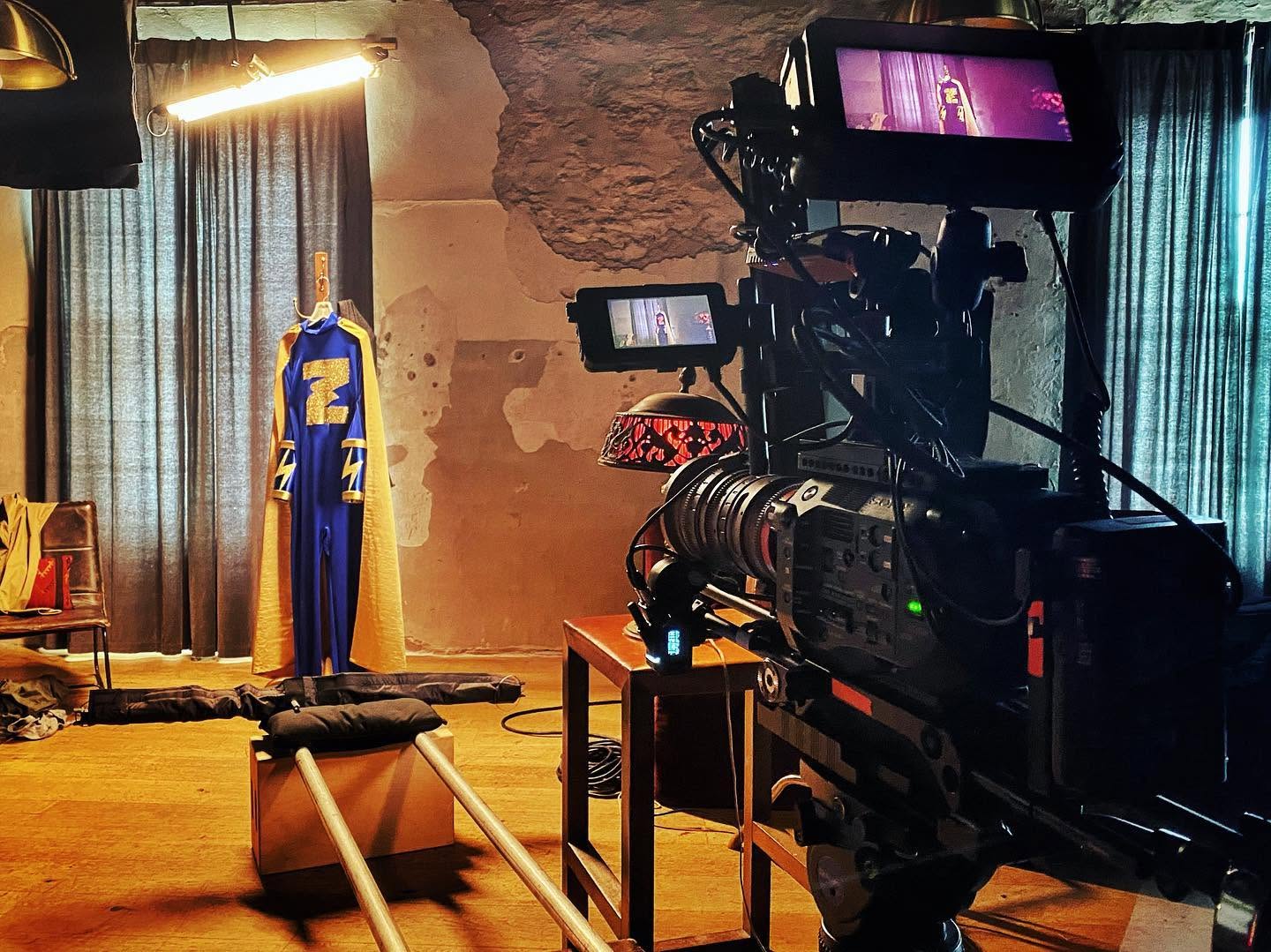 Camera setup in a room with curtains, a mannequin in a blue and yellow outfit, and a stool with a black cushion.