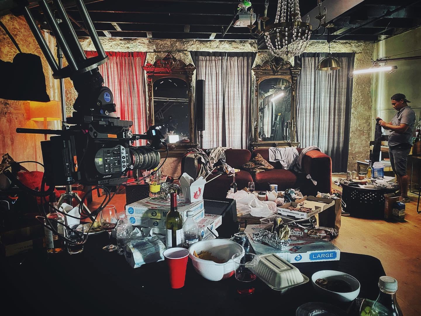 Living room with sofa, curtains, and chandelier, cluttered with bottles, cups, and papers, with a person standing on the right side.