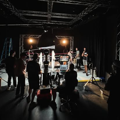People in a studio with lighting and camera equipment, preparing for a photoshoot or filming, with a black backdrop and stage lights.