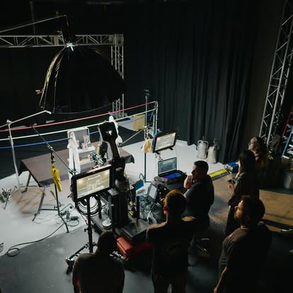 People working on a film or TV set with cameras, monitors, and lighting equipment, in a dark studio with a black curtain backdrop.