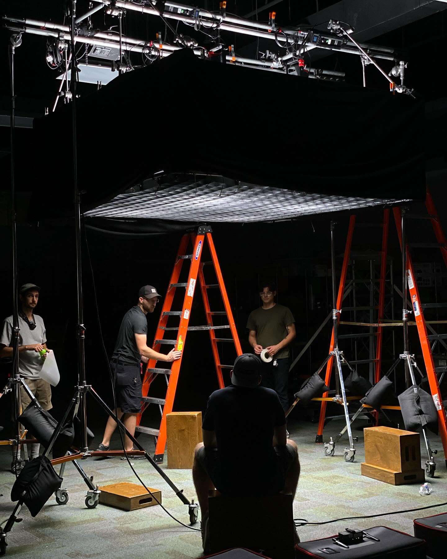 People working on a stage setup with ladders, lighting equipment, and a large black fabric overhead.