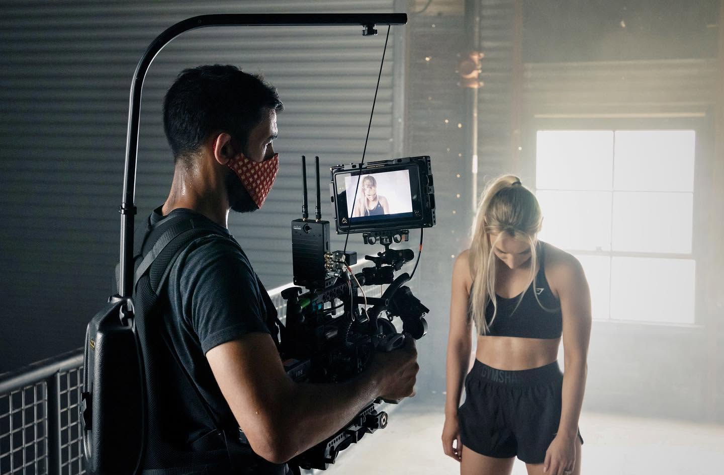 A person operating a camera records a woman in a dark sports outfit in a dimly lit room with a bright window.