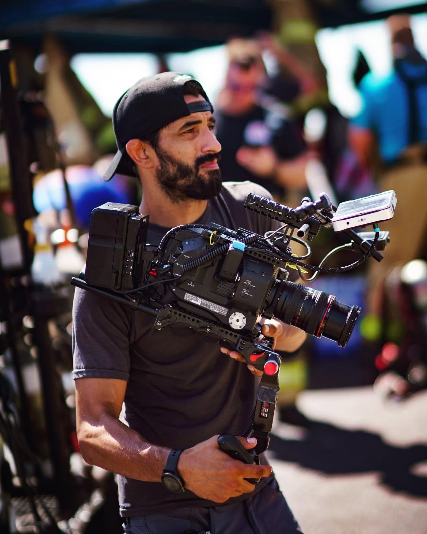 Man with a beard and cap holding a professional camera rig outdoors, with people in the background.