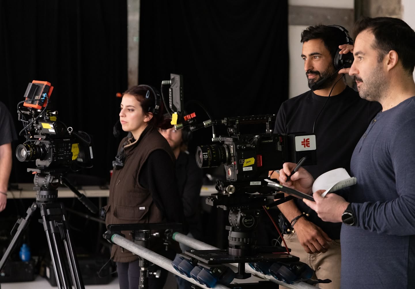 Film crew members operate cameras and equipment in a studio setting, with a woman and two men present.
