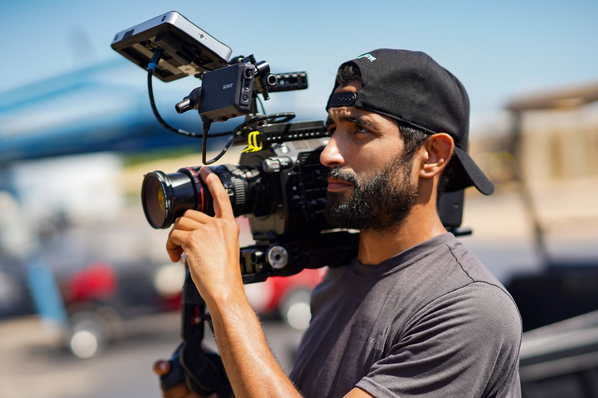 Man with a beard wearing a black cap and gray shirt holding a professional video camera outdoors.