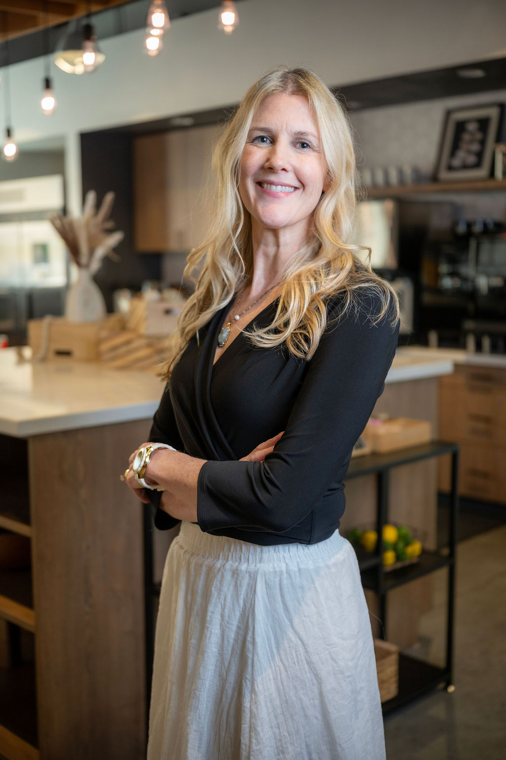 Woman with long blonde hair smiling, standing in a modern kitchen with wooden cabinets and hanging lights.