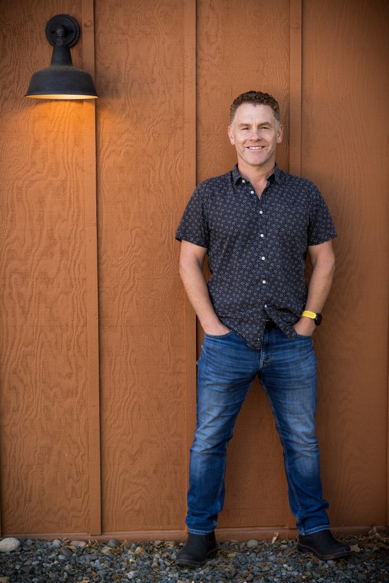 Man standing with hands in pockets, smiling, in front of wooden wall with a wall-mounted lamp on the left.