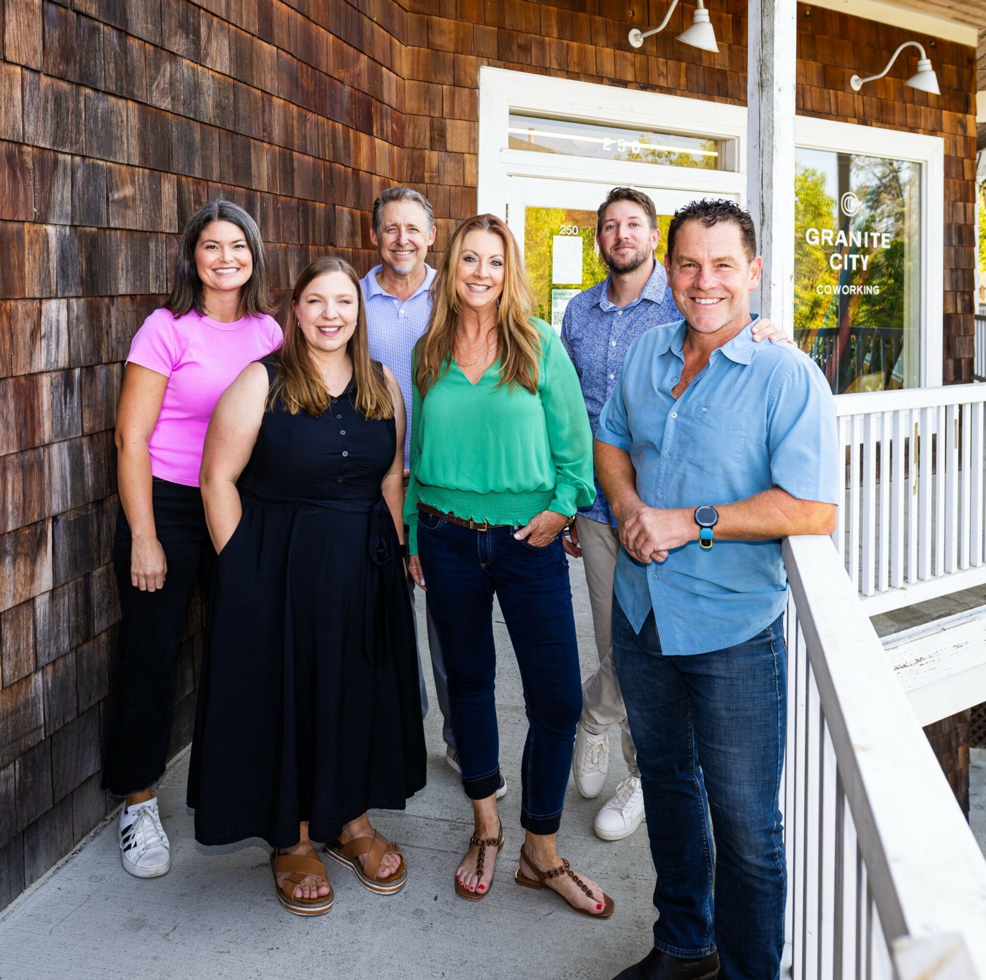 Group of six people standing outside a building, smiling, with a wooden wall and window behind them.