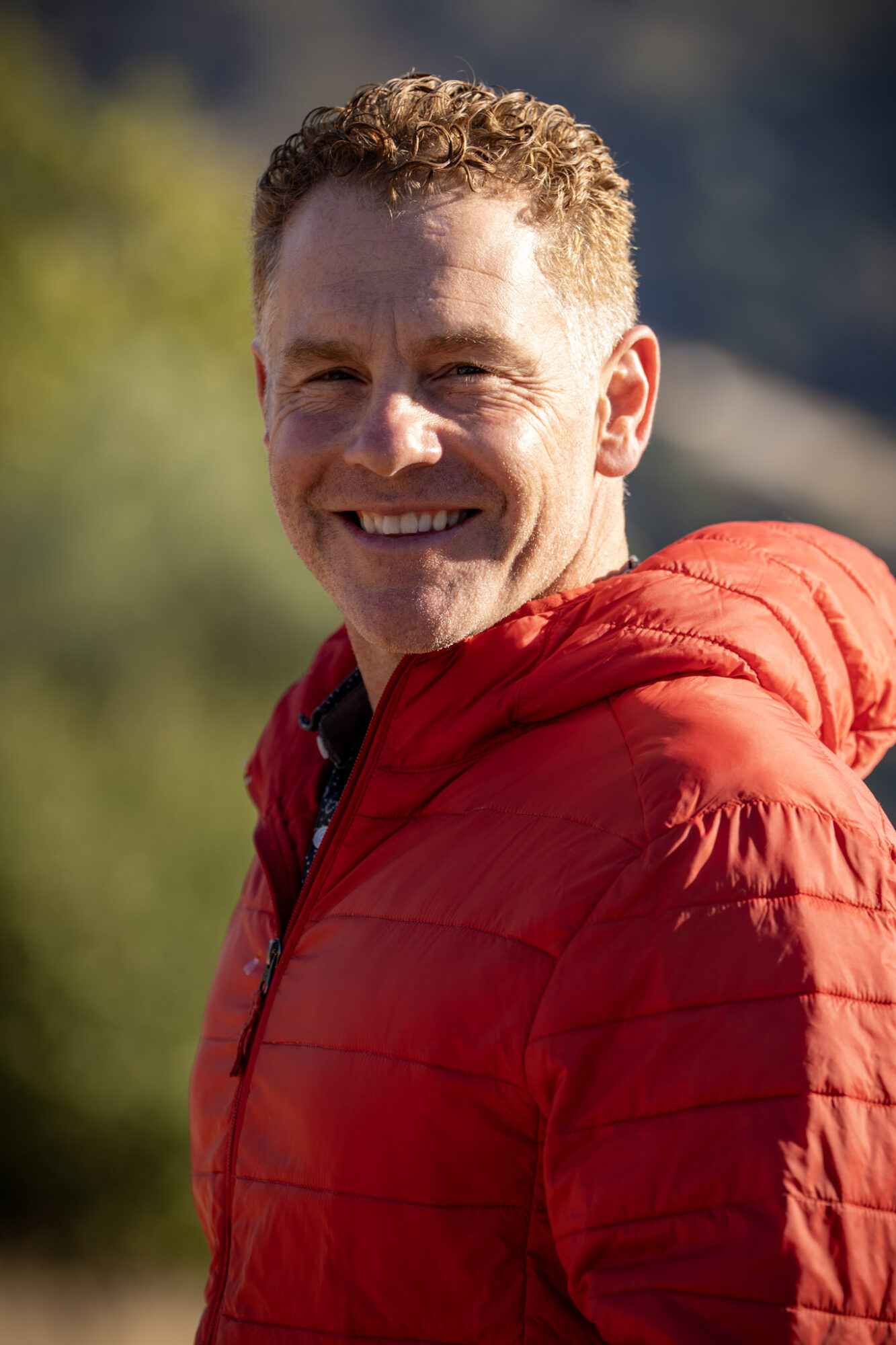 Smiling man with curly hair wearing a red jacket outdoors, background blurred with greenery and mountains.