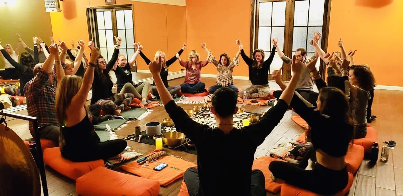 Group of people participating in a yoga or meditation session in a brightly lit room with orange walls and large windows, sitting on cushions with raised arms.