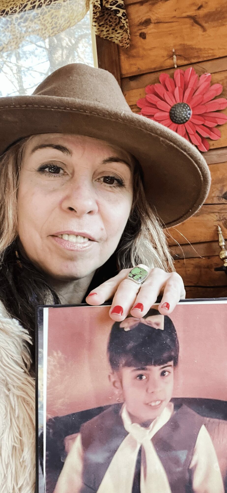 Woman wearing a hat holding a photo of a young boy, wooden wall and flower decoration in background.