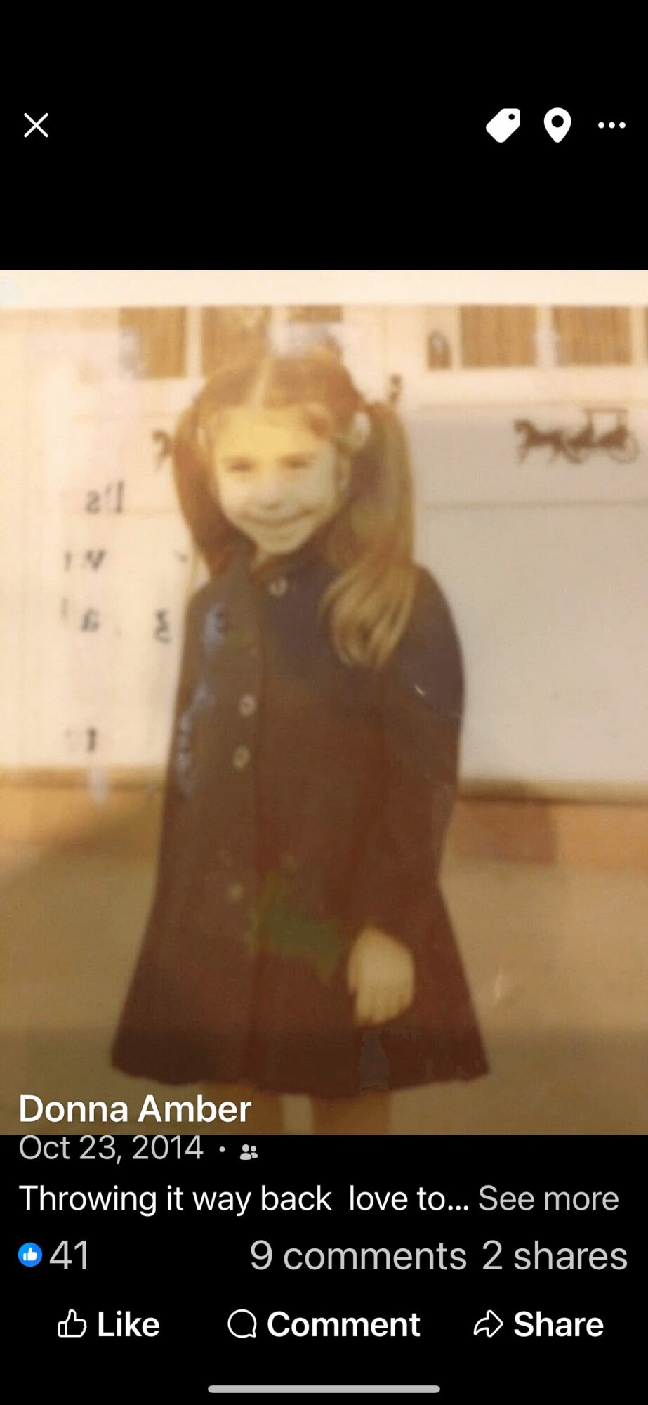 Child with pigtails wearing a dark coat, standing outdoors in front of a building, smiling.