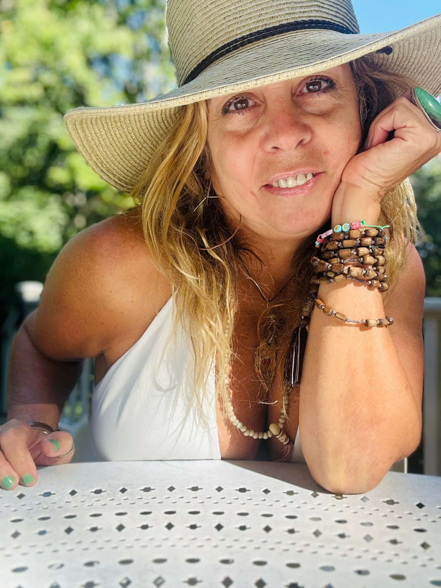 Woman wearing a large sun hat, smiling, resting her chin on her hand, outdoors with greenery in background.