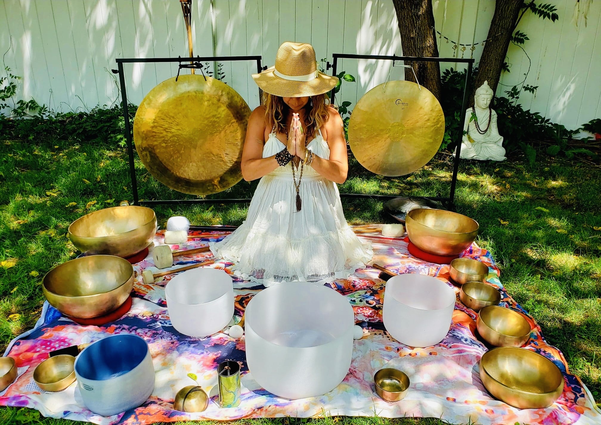 Person in white dress and hat sitting on colorful cloth surrounded by singing bowls and large hanging gongs outdoors.