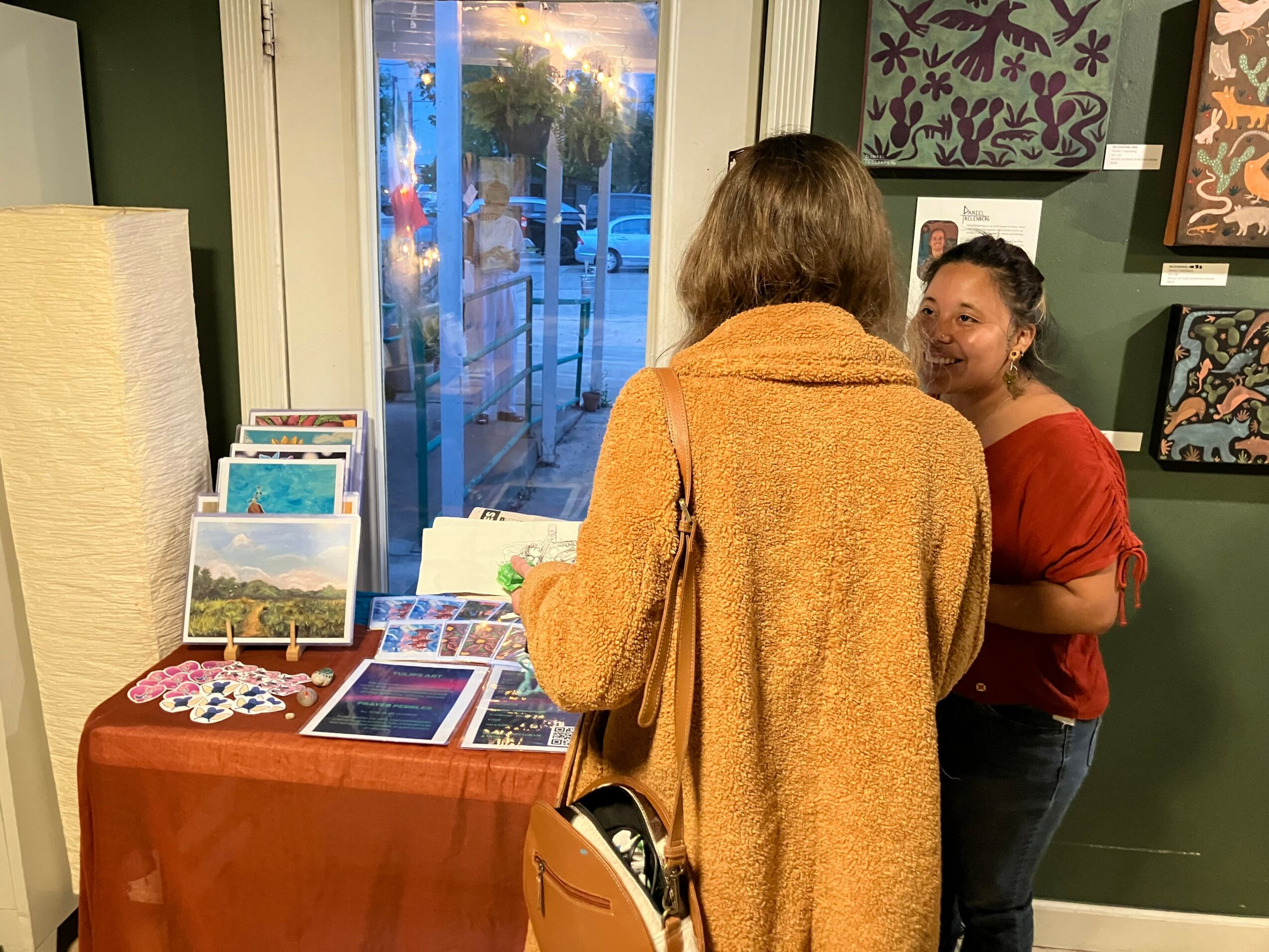 Two women talking inside a room with artwork on the wall, one facing away, the other smiling, near a table with brochures and a tablet.