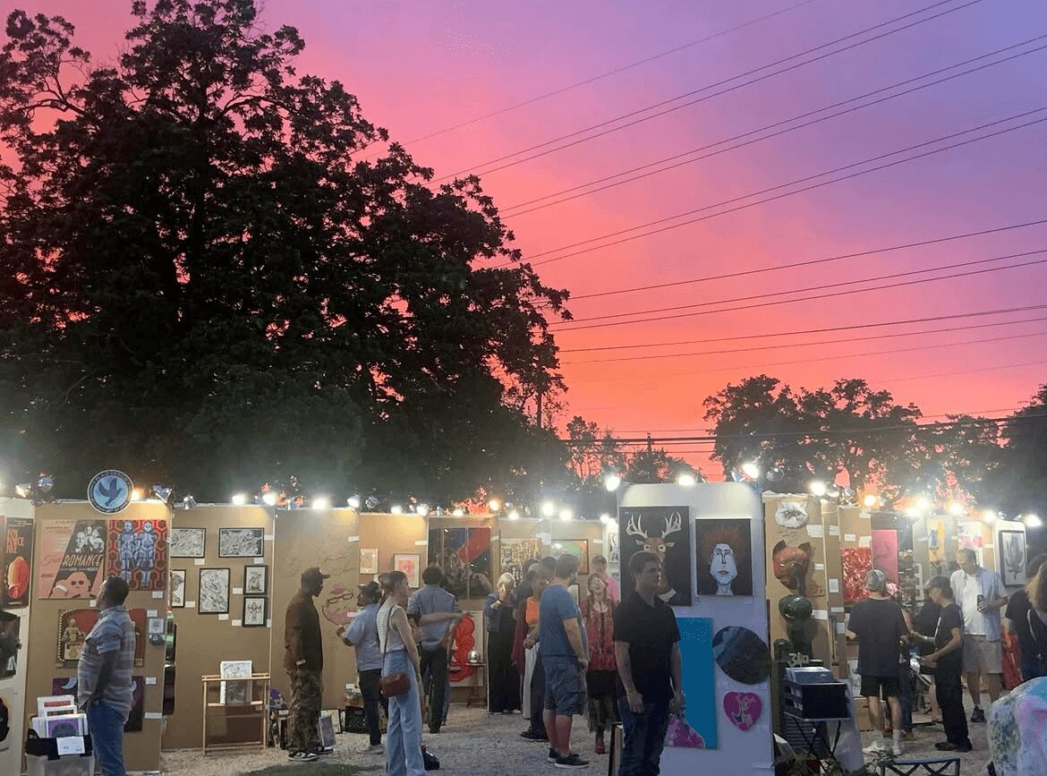 People browsing art and photo displays outdoors during sunset with colorful sky and trees in background.