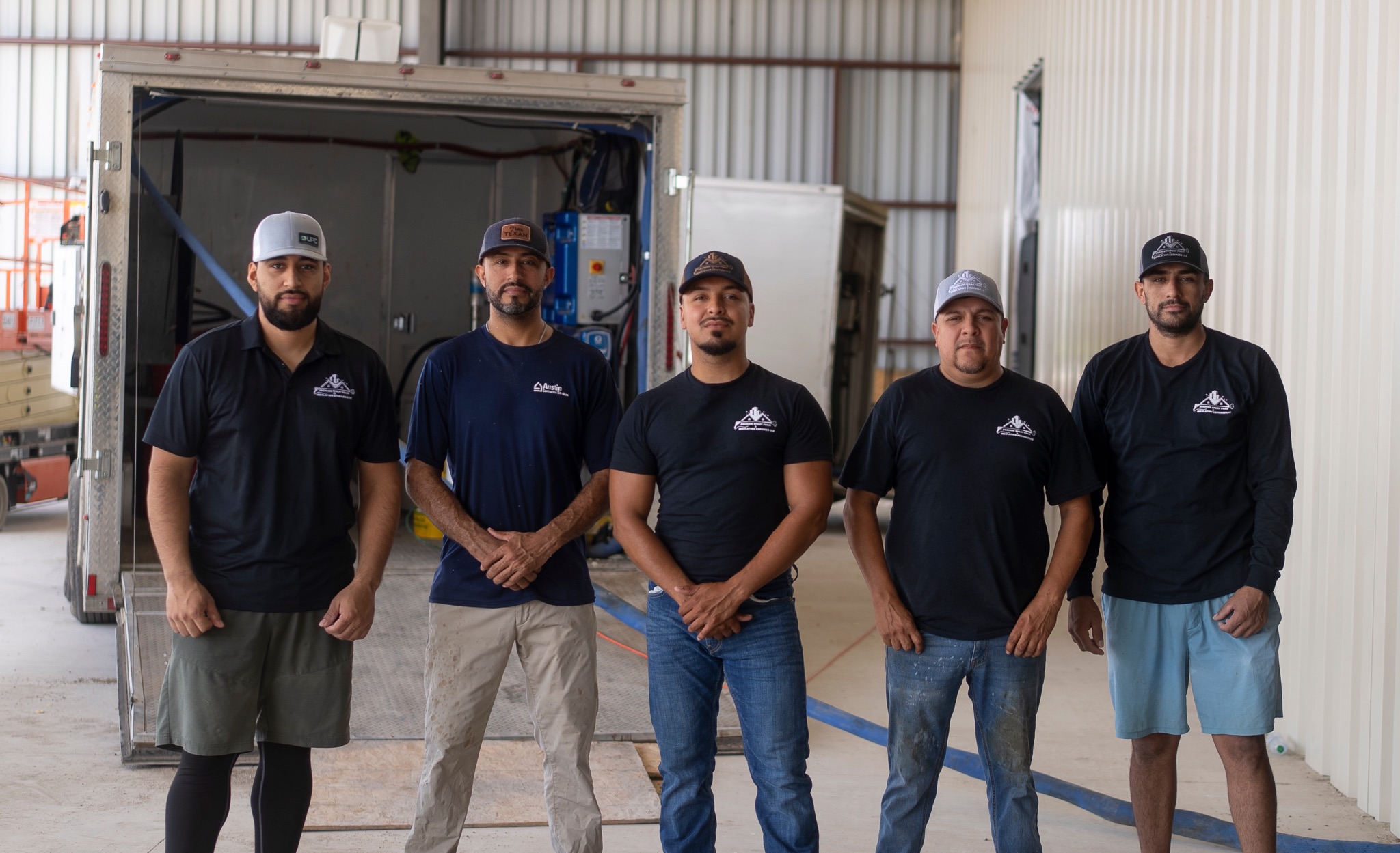 Five men stand in front of a truck, wearing black shirts and caps, with a warehouse background.