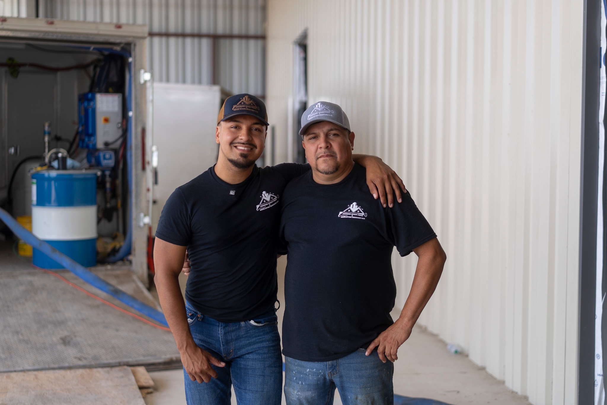 Two men stand side by side, one with arm around the other's shoulder, inside a warehouse or industrial space.