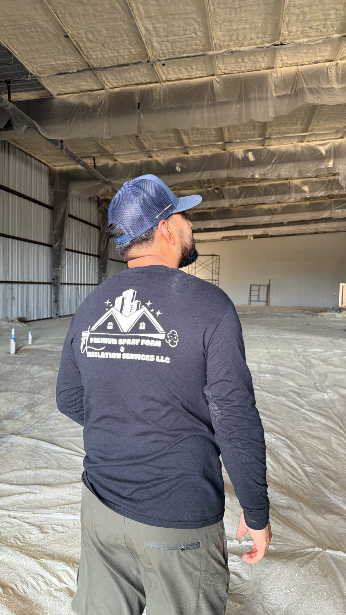 Person wearing a blue helmet and dark jacket with a graphic on the back, standing inside a large industrial space.