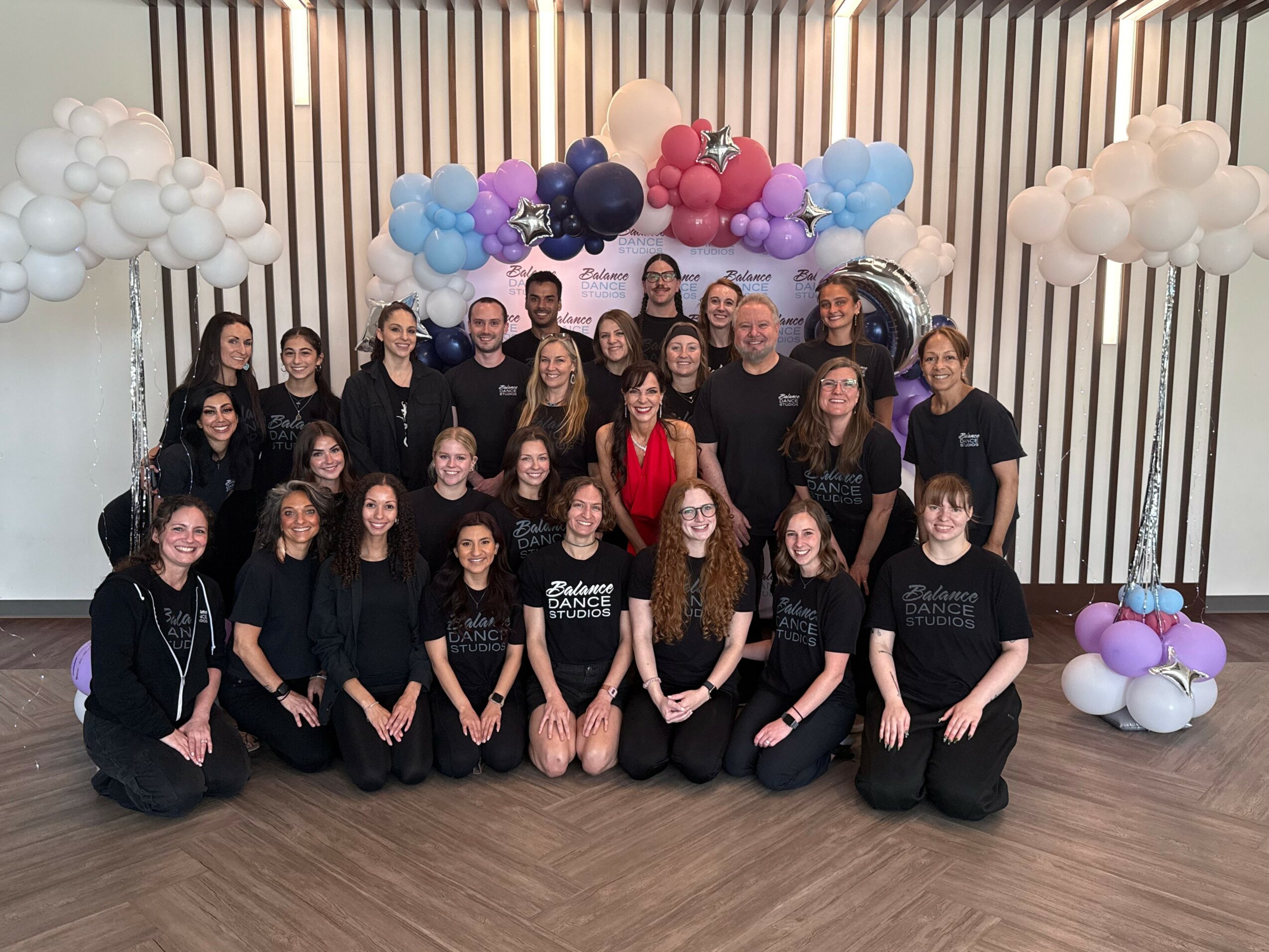 Group of people posing in front of colorful balloon decorations, some wearing black shirts, indoors with wooden floor and striped wall.