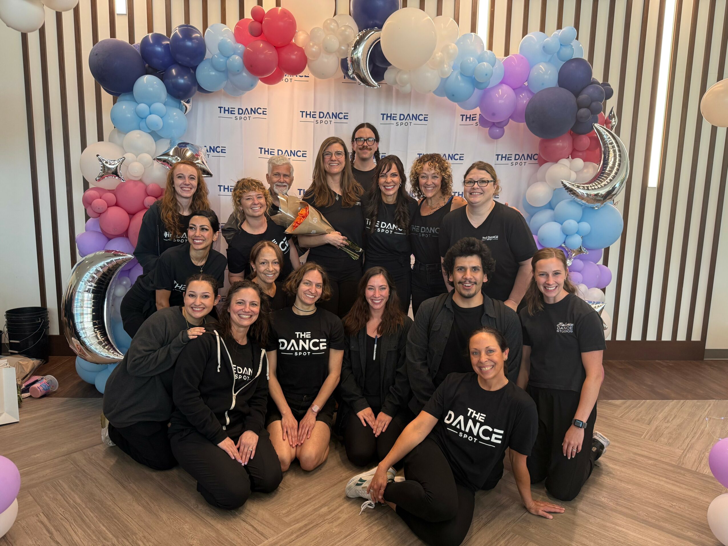 Group of people posing in front of balloon arch with stars and moon decorations, indoors, smiling, some wearing matching shirts.