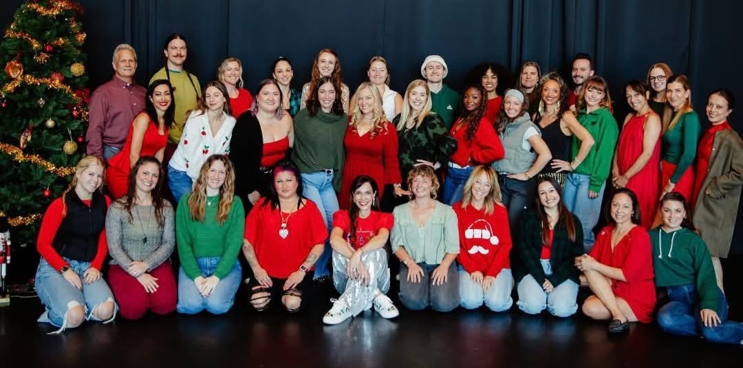 Group of people posing on stage, some wearing holiday-themed clothing, with a Christmas tree on the left.