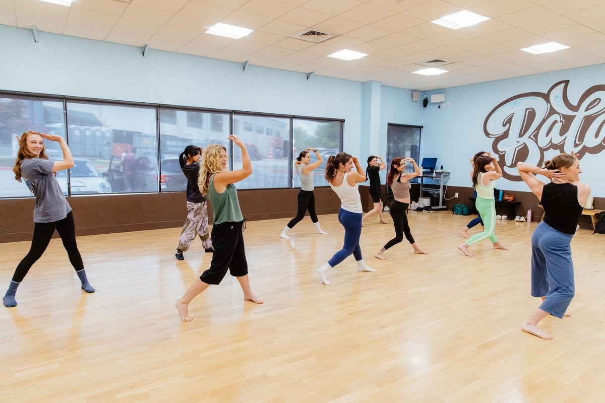 Group of people practicing dance moves in a spacious studio with large windows and a logo on the wall.