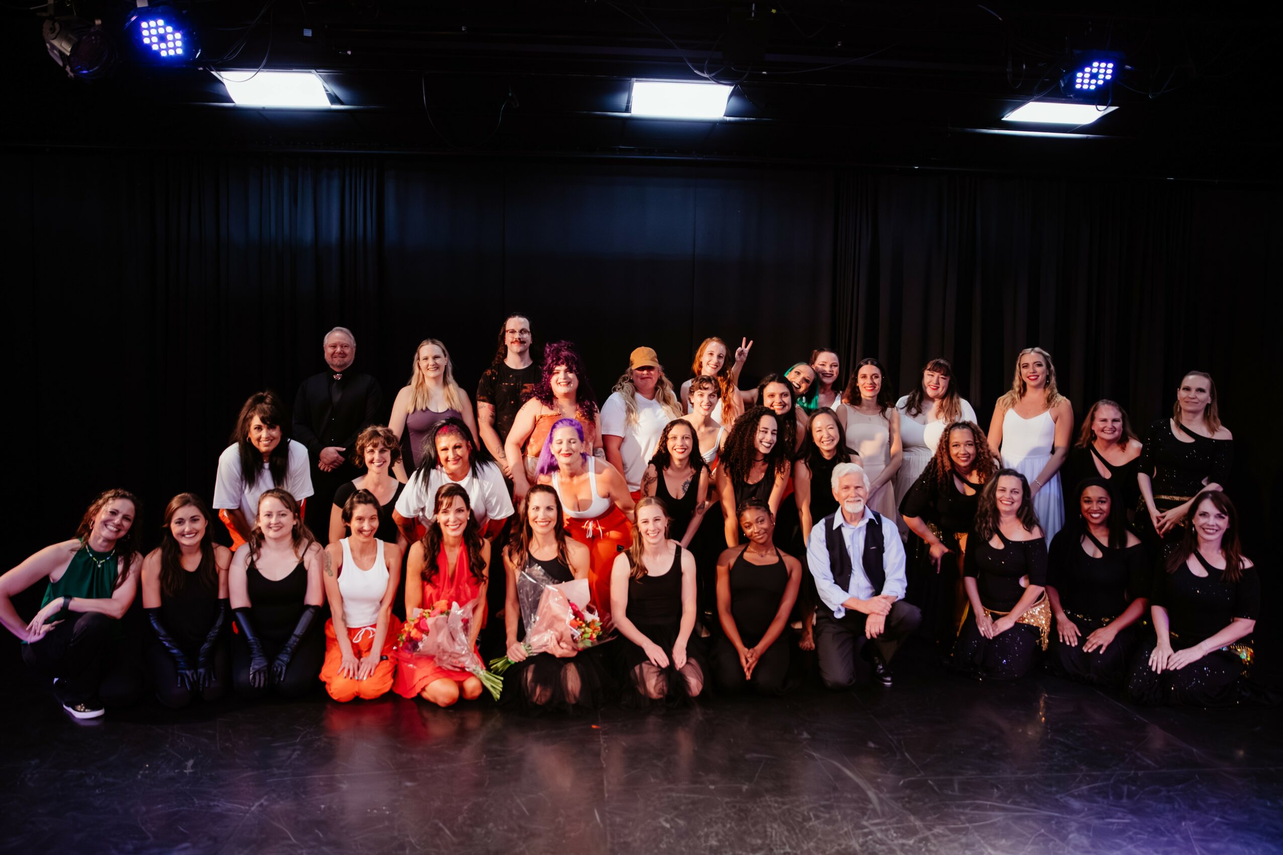 Group of people on stage, some sitting and some standing, with a black curtain background, stage lights overhead.