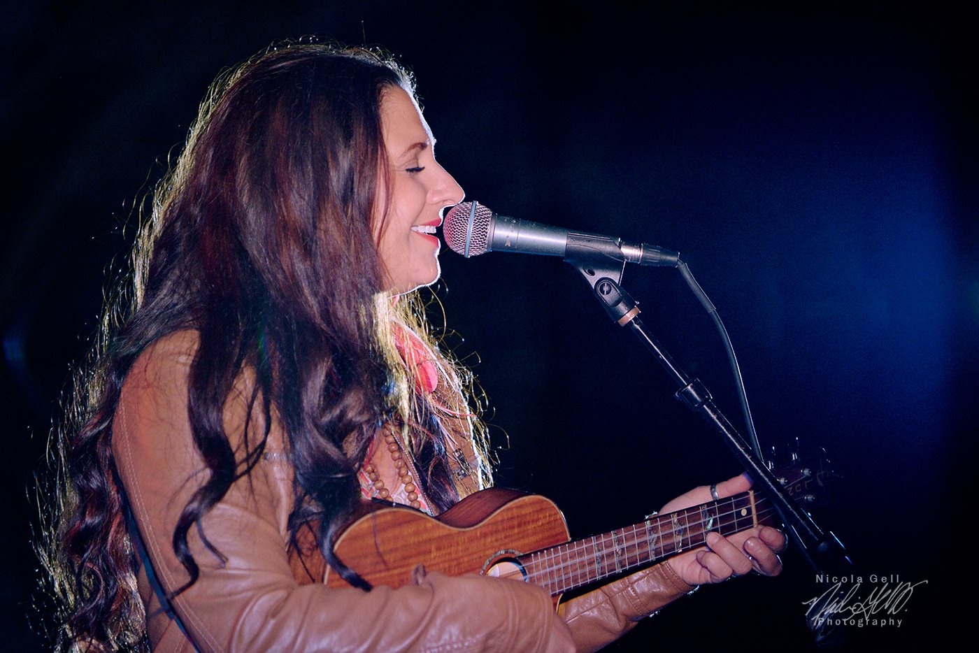 Woman singing into microphone while playing ukulele on stage, dark background, stage lighting.
