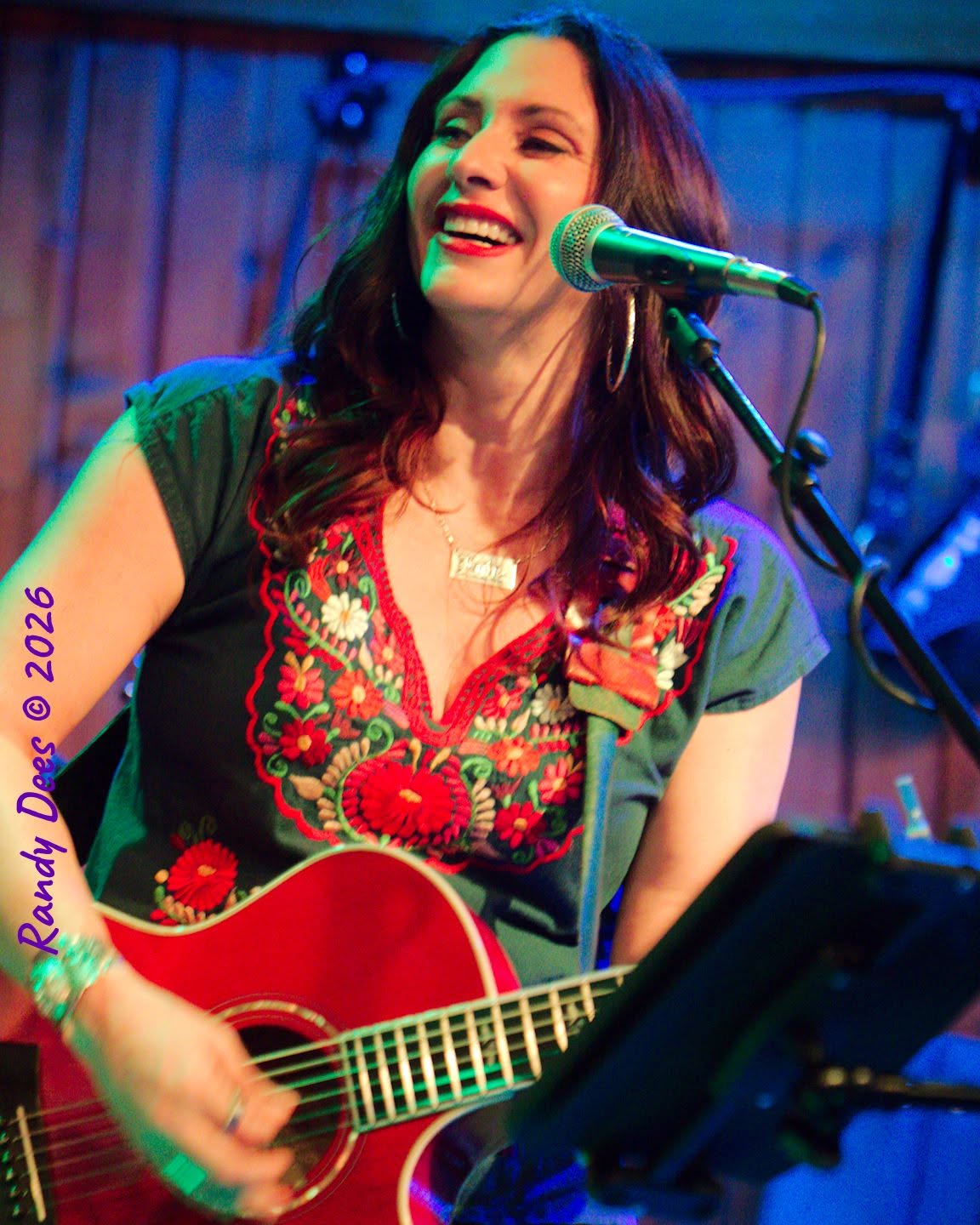 Woman singing into a microphone while playing a red guitar, smiling, with a wooden background.