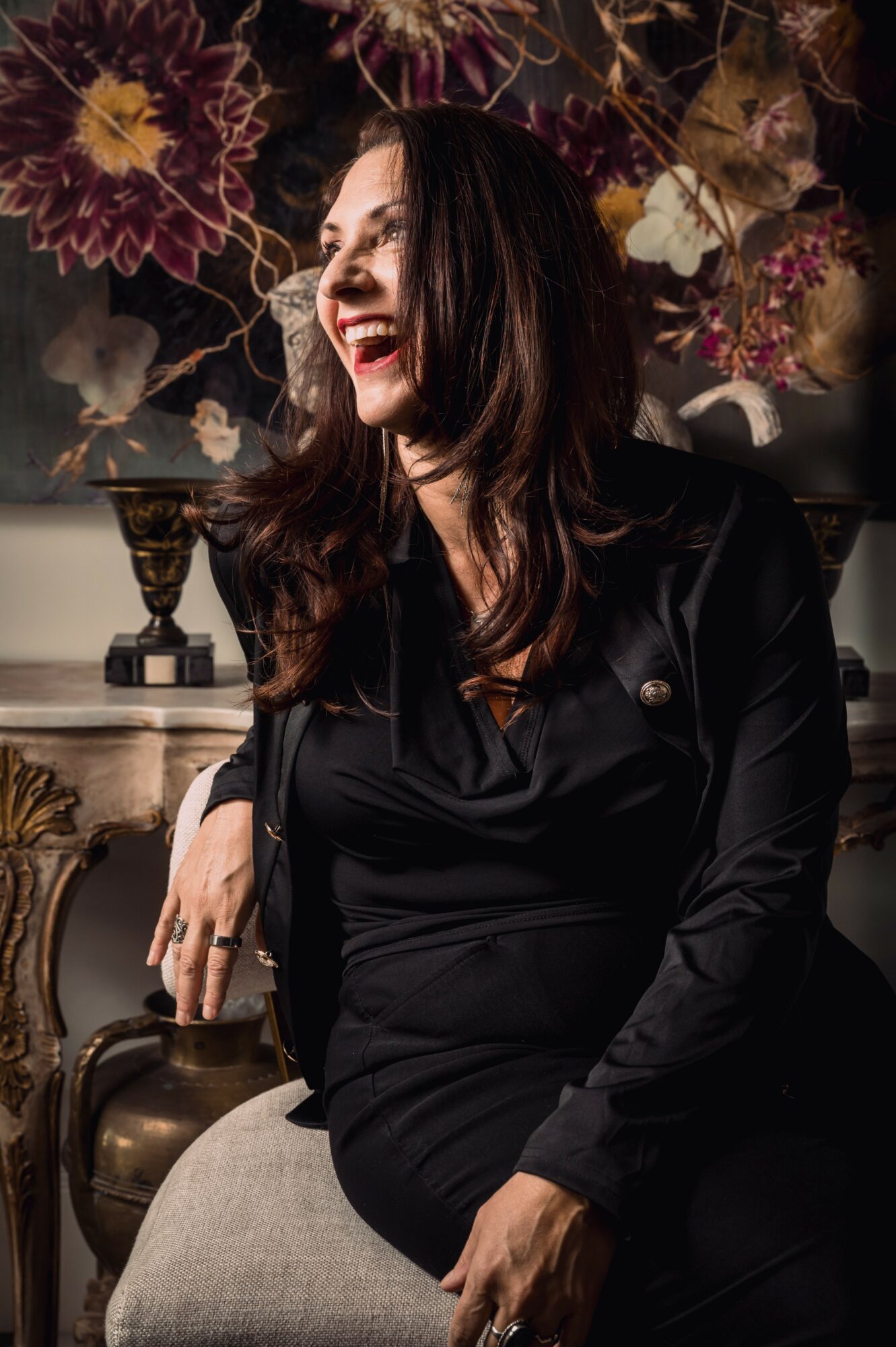 Woman with dark hair smiling, sitting in front of floral wallpaper, wearing a black outfit, with a decorative background.