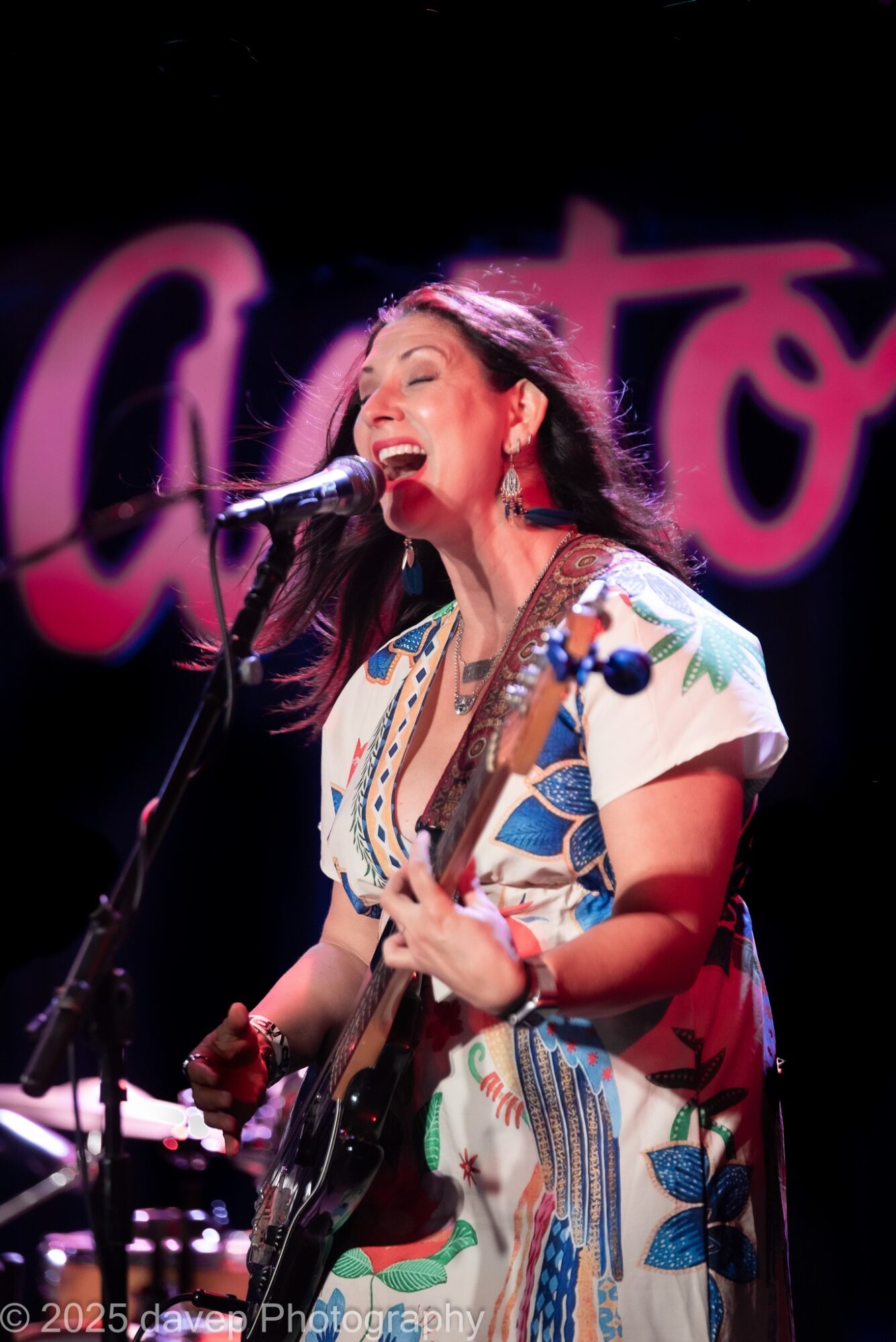 Woman singing and playing guitar on stage with a microphone, wearing a colorful dress, with a pink and purple background.