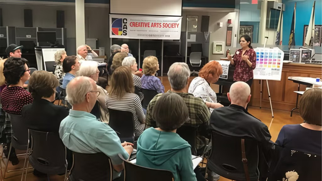 People seated in a room listening to a speaker at a Creative Arts Society event, with a banner and colorful chart visible.