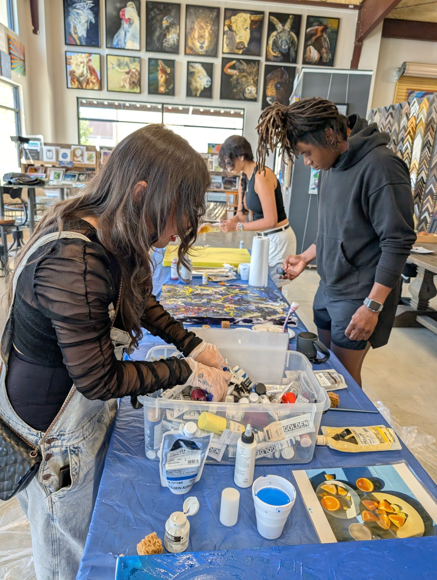 People working on art projects at a table in an art studio, with paintings displayed on the wall behind them.