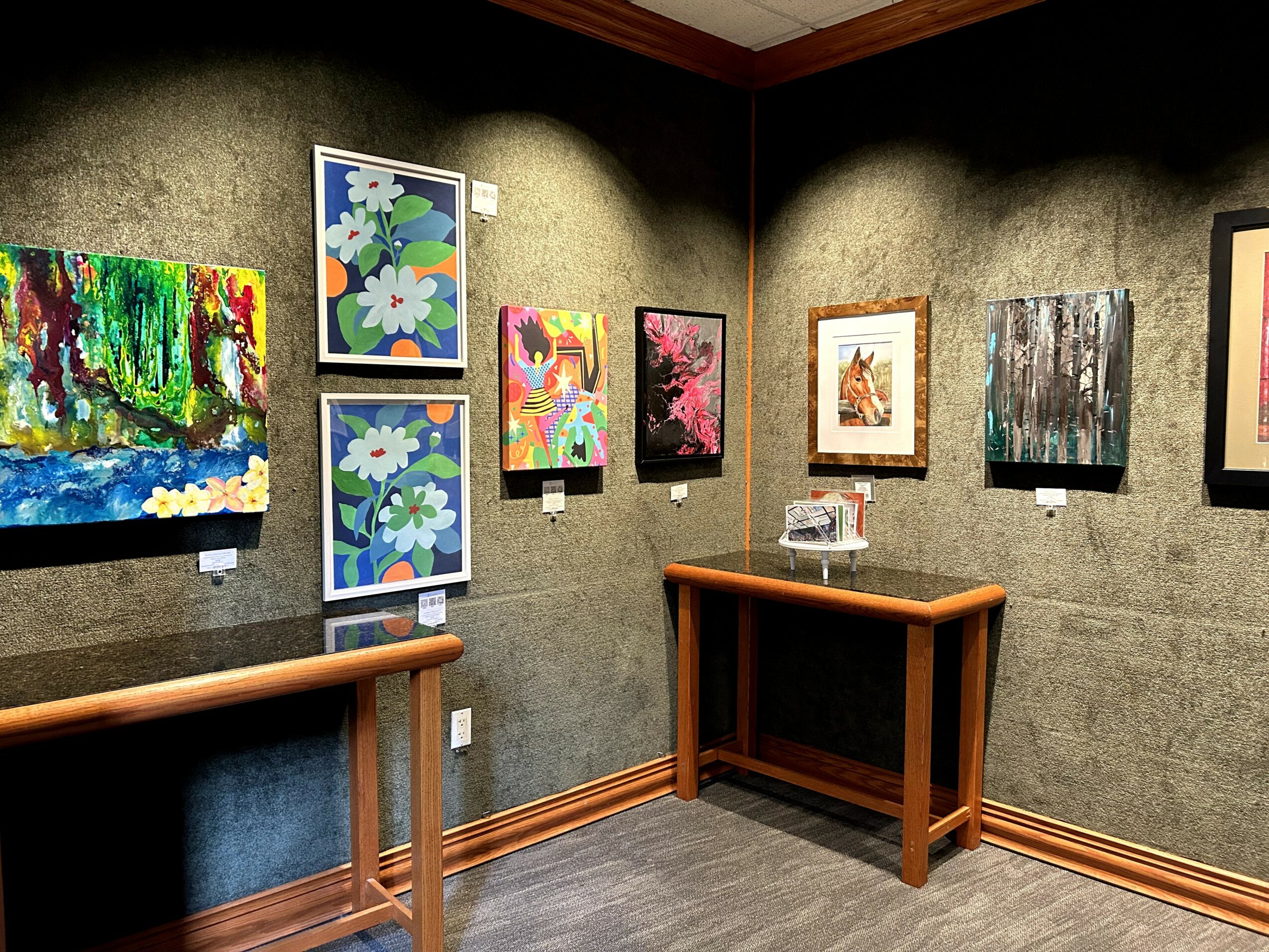 Gallery wall with colorful paintings, some framed, displayed on dark textured walls, with wooden furniture and gray flooring.