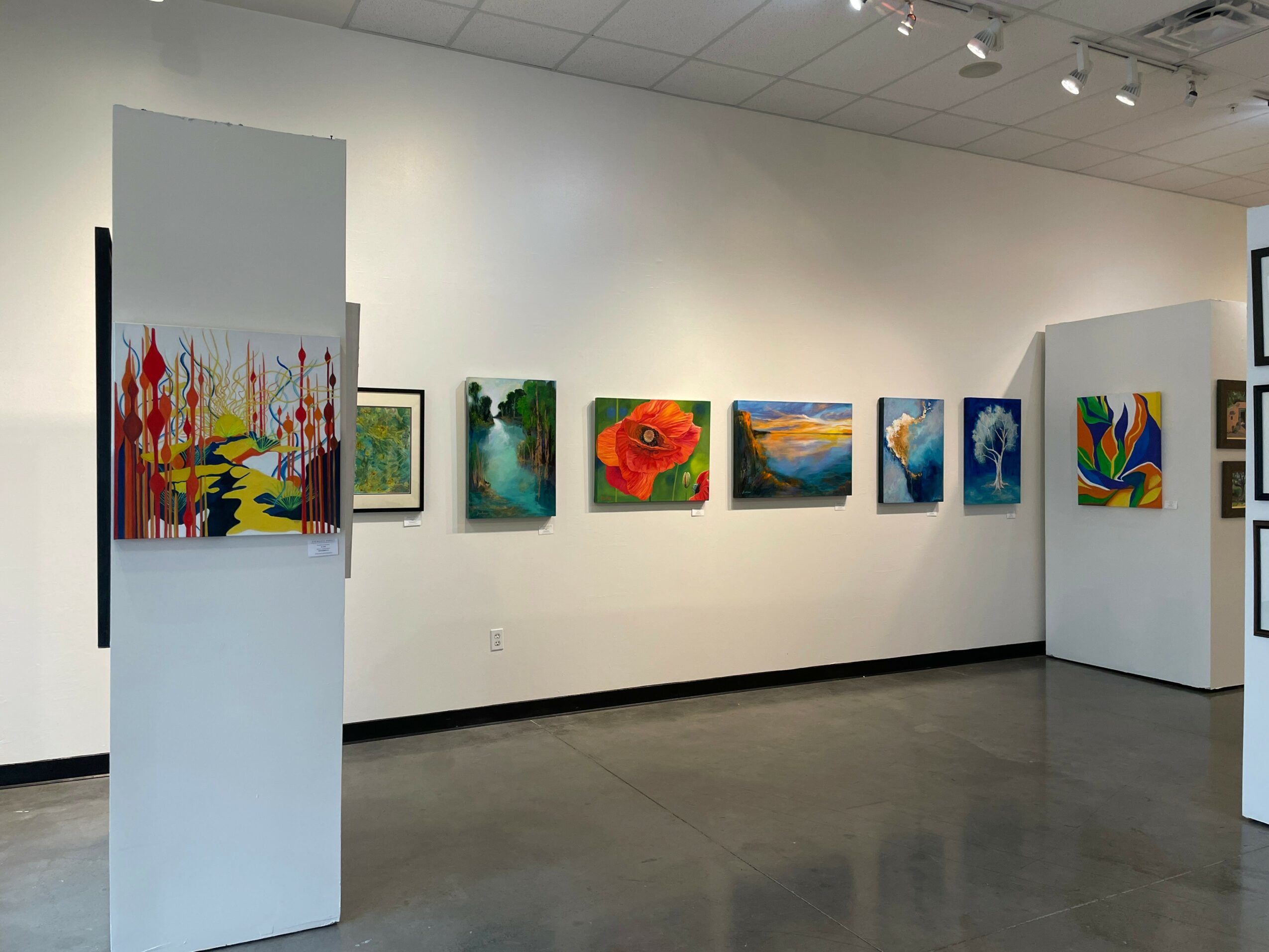 Gallery with colorful paintings on white walls, some on display on white pedestals, in a well-lit room.