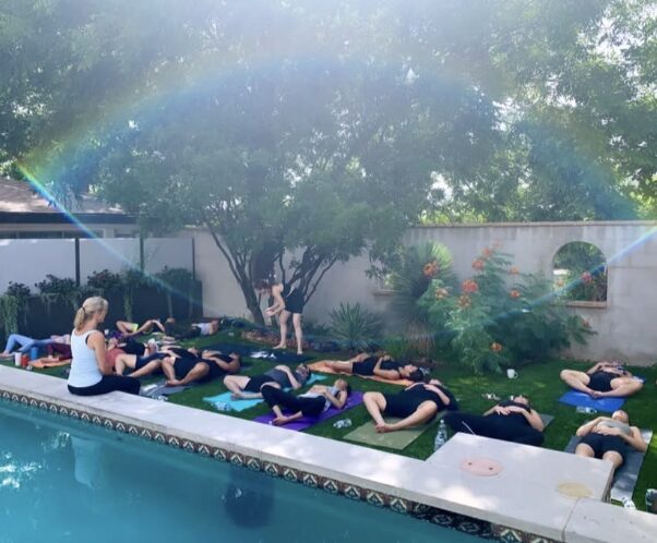 Group of people practicing yoga outdoors on mats beside a pool, with a woman instructor and a rainbow in the sky.
