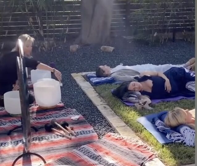 People lying on mats outdoors, with one person sitting nearby, in a garden setting with trees and a fence.